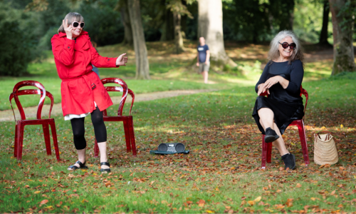 2 Frauen im Park. Eine Frau telefoniert im stehen, die andere sitzt am Stuhl und lacht