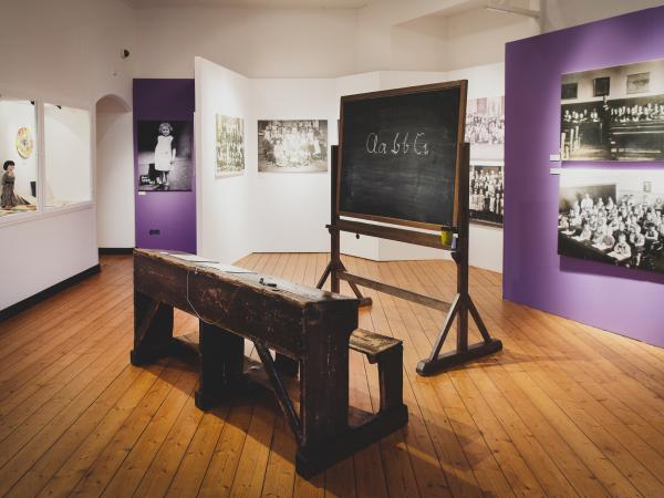Blick in einen Ausstellungsraum mit Fotos, einer Schulbank und einer Tafel
