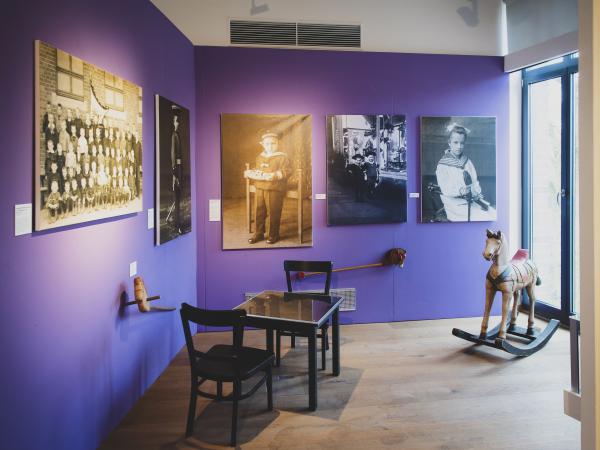 Blick in einen Ausstellungsraum mit einem Tisch, 2 Stühlen, 1 Schaukelpferd und Fotos an den Wänden