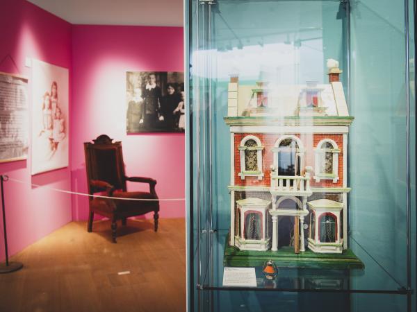 Blick in einen Ausstellungsraum mit einem Stuhl und einem Puppenhaus in einer Vitrine