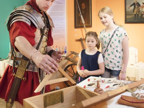 Ein Mann in römischer Rüstung zeigt zwei Mädchen etwas.